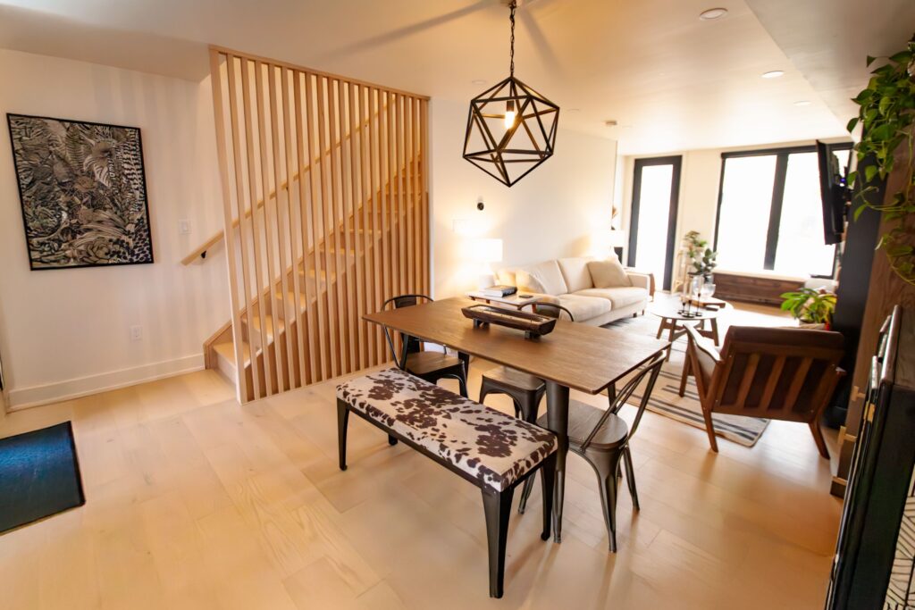 Modern open-concept living and dining area with a wooden table, bench, armchairs, geometric pendant light, light wood floors, and a staircase with vertical wooden slats. Natural light fills the space.