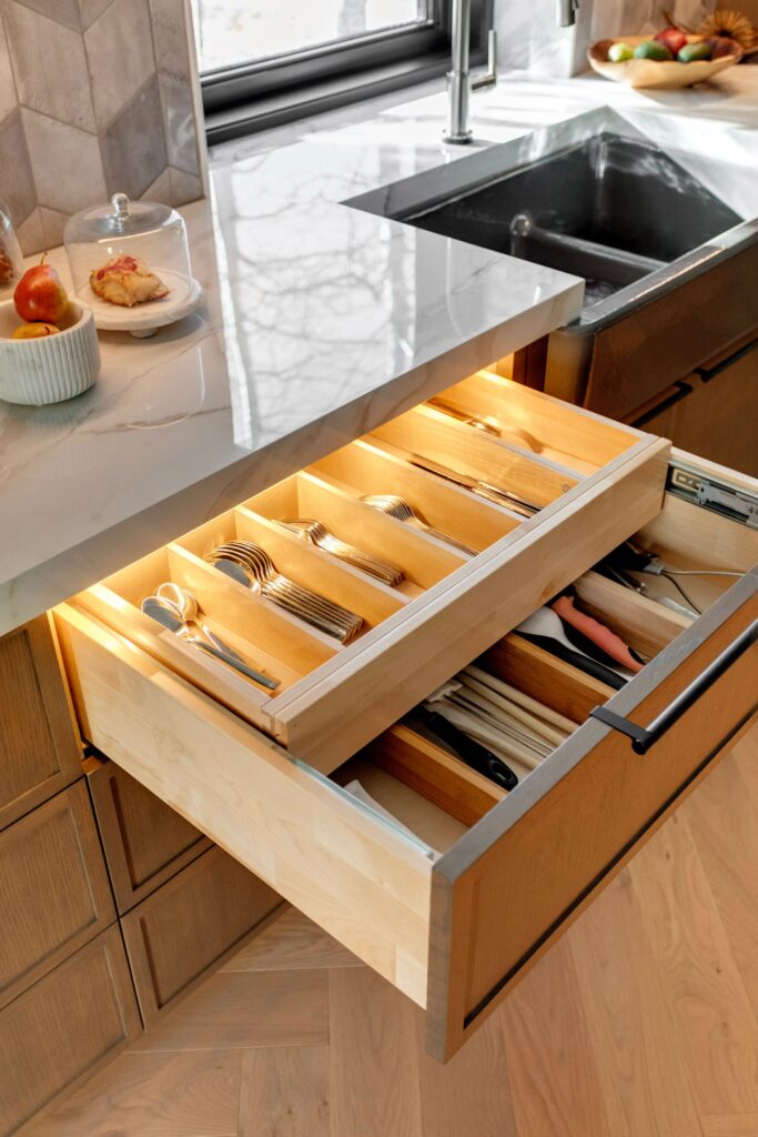 A kitchen drawer beneath a marble countertop is open, revealing organized compartments with utensils, including forks, knives, spoons, and scissors. Soft lighting illuminates the drawer. A sink and food items are visible on the counter.