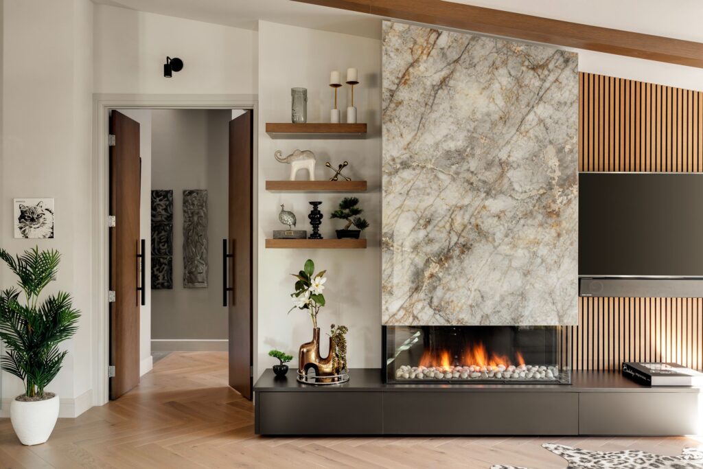 A modern living room with a large marble fireplace, decorative shelves, a potted plant, a TV mounted on a wood slat wall, and an open doorway leading to another room. The decor is neutral with gold and black accents.