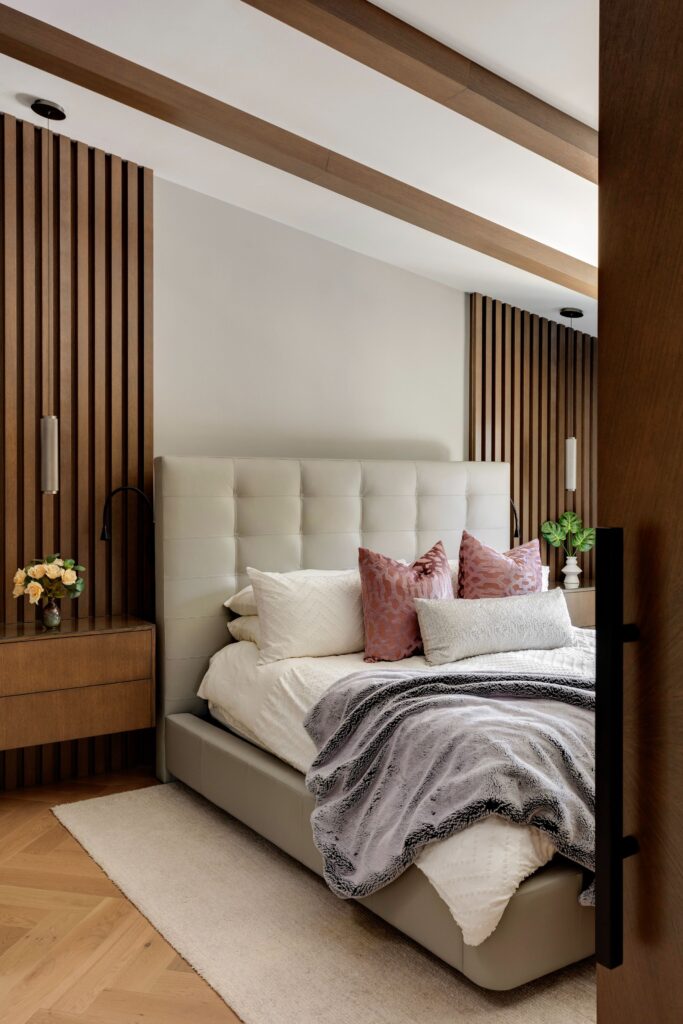 A modern bedroom with a tufted light-gray bed, white bedding, purple accent pillows, wood-paneled walls, two bedside tables with flowers, and soft lighting. The color palette is neutral with warm wood accents.