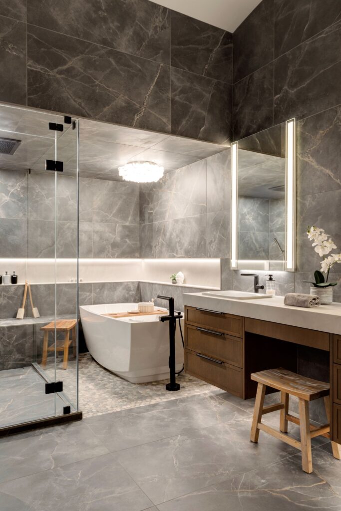 Modern bathroom with gray marble walls and floor, a freestanding white bathtub, glass shower enclosure, wooden vanity with large mirror, lighted shelves, and a wooden stool. An orchid plant sits on the counter.