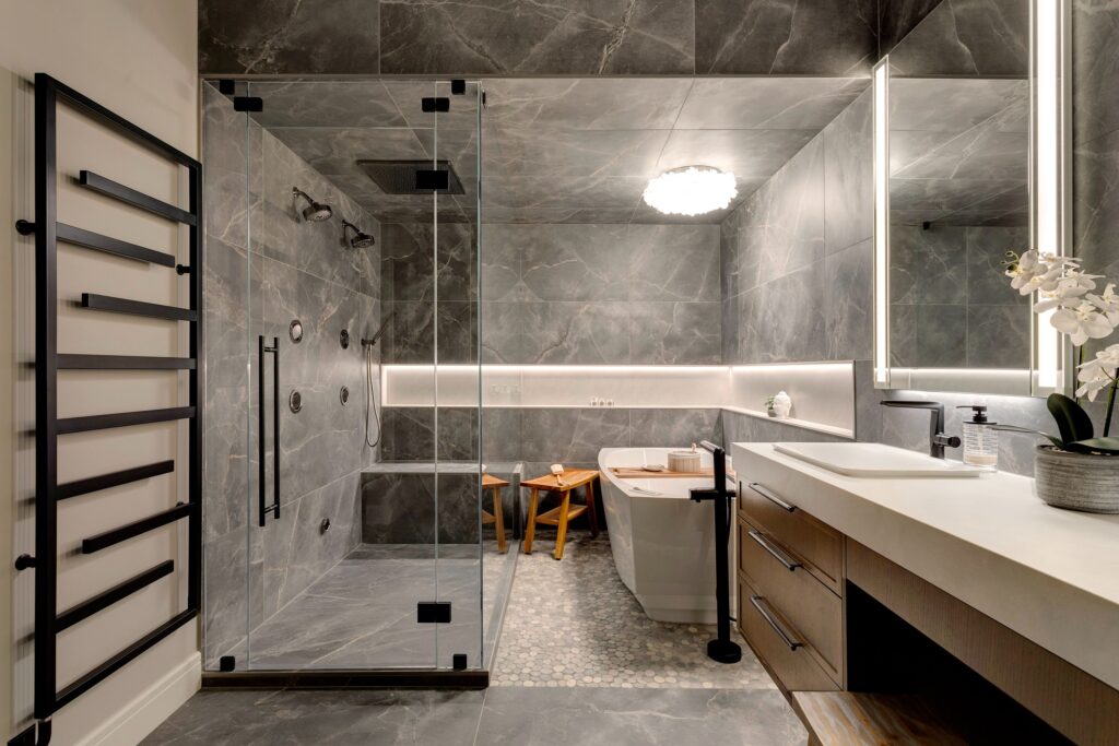 Modern bathroom with gray marble walls and floor, glass shower enclosure, soaking tub, large mirror with LED lighting, wooden vanity, heated towel rack, and a white orchid on the counter.