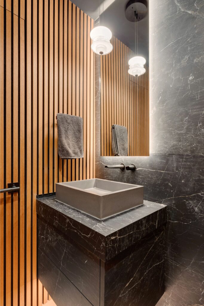 Modern bathroom with wood slat walls, a rectangular stone sink on a black marble vanity, a large mirror, wall-mounted faucet, and two hanging pendant lights with frosted glass shades.
