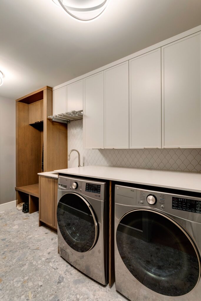 A modern laundry room features front-loading washer and dryer units, white upper cabinets, a built-in wooden shelving unit, a sink, and light gray patterned flooring.