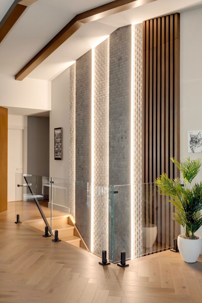 Modern staircase with glass railing and wooden handrail, featuring a dramatic wall with vertical wooden slats, illuminated textured panels, and decorative indoor plant; artwork hangs on the light-colored walls.