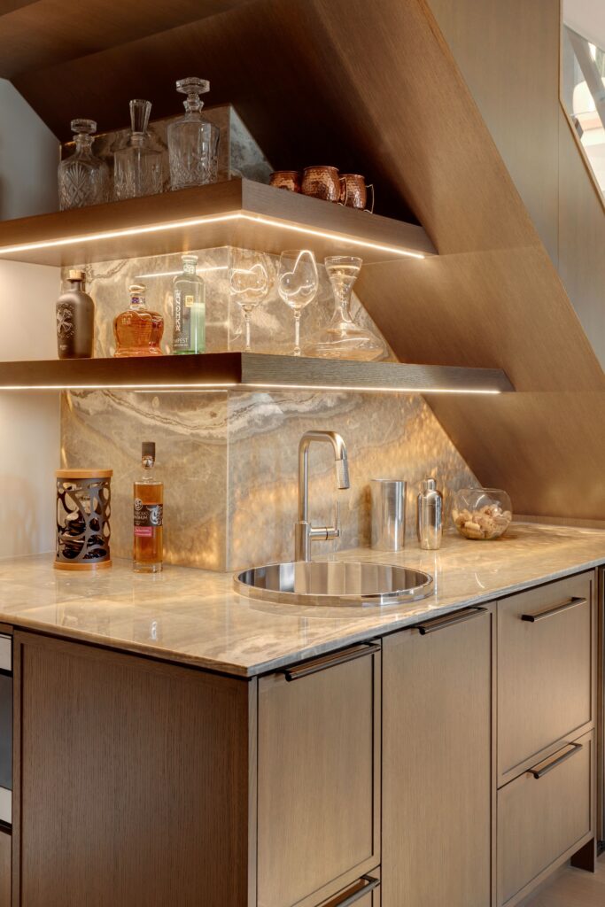 A modern home bar with wooden cabinets, marble countertop and backsplash, a round sink, glassware, decanters, and liquor bottles on illuminated shelves under a staircase.