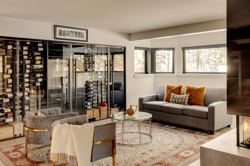 A modern living room with a glass-enclosed wine cellar, gray sofa with orange pillows, two gray armchairs, a round glass coffee table, and a patterned rug. Sunlight streams in through wide windows.
