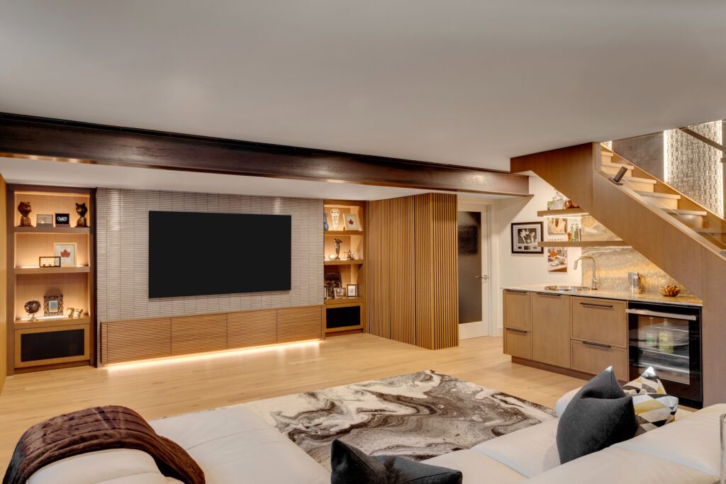 A modern living room with a large wall-mounted TV, built-in shelves displaying decor, a small kitchenette under wooden stairs, and a white sectional sofa with a patterned rug on light wood flooring.