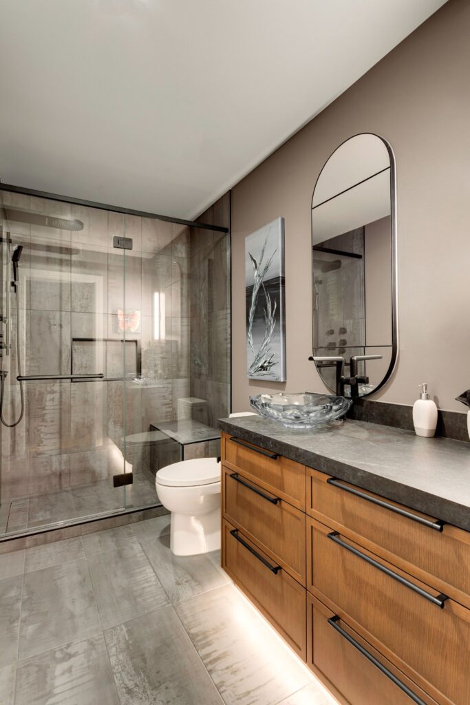 Modern bathroom with a glass-enclosed shower, wooden vanity with black handles, oval mirror, vessel sink, toilet, wall art, and neutral-colored walls and floor tiles.