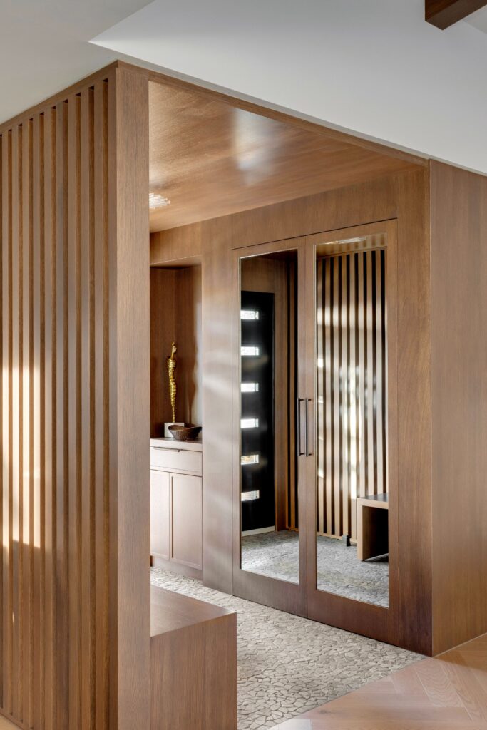A modern walk-in closet with wood panel walls, a large floor-to-ceiling mirror on a closet door, patterned grey carpeting, and ambient light from a dressing table with illuminated shelves.
