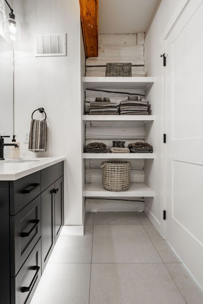 Open shelving in a modern bathroom displays neatly folded gray towels and woven baskets. The black vanity has a white countertop, and the wall is painted white with wood accents. The space is clean and organized.