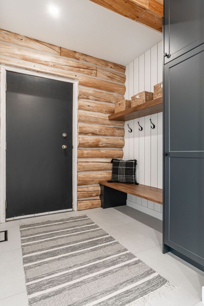 A mudroom with log cabin walls, a black door, striped rug, bench with a plaid pillow, wooden shelves with baskets, and coat hooks on a white shiplap wall.
