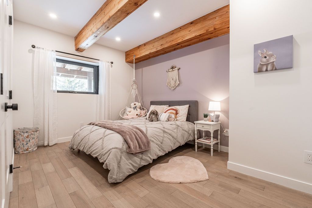 A cozy bedroom with wood beams, a bed with plush pillows and stuffed animals, a mauve accent wall, a small nightstand with a lamp, sheer white curtains, and a heart-shaped rug on a wooden floor.