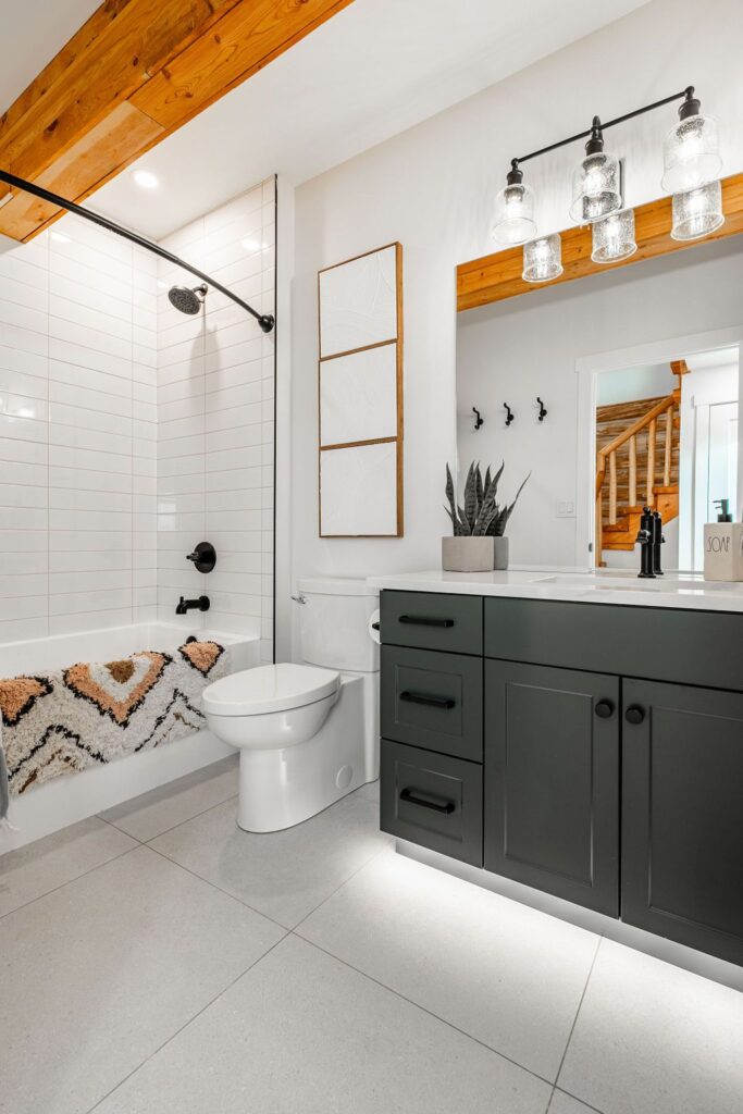 Modern bathroom with white tile shower, black fixtures, a patterned bath mat, dark vanity with a white countertop, a plant, large mirror, and exposed wood beams on the ceiling. Warm lighting creates a cozy atmosphere.