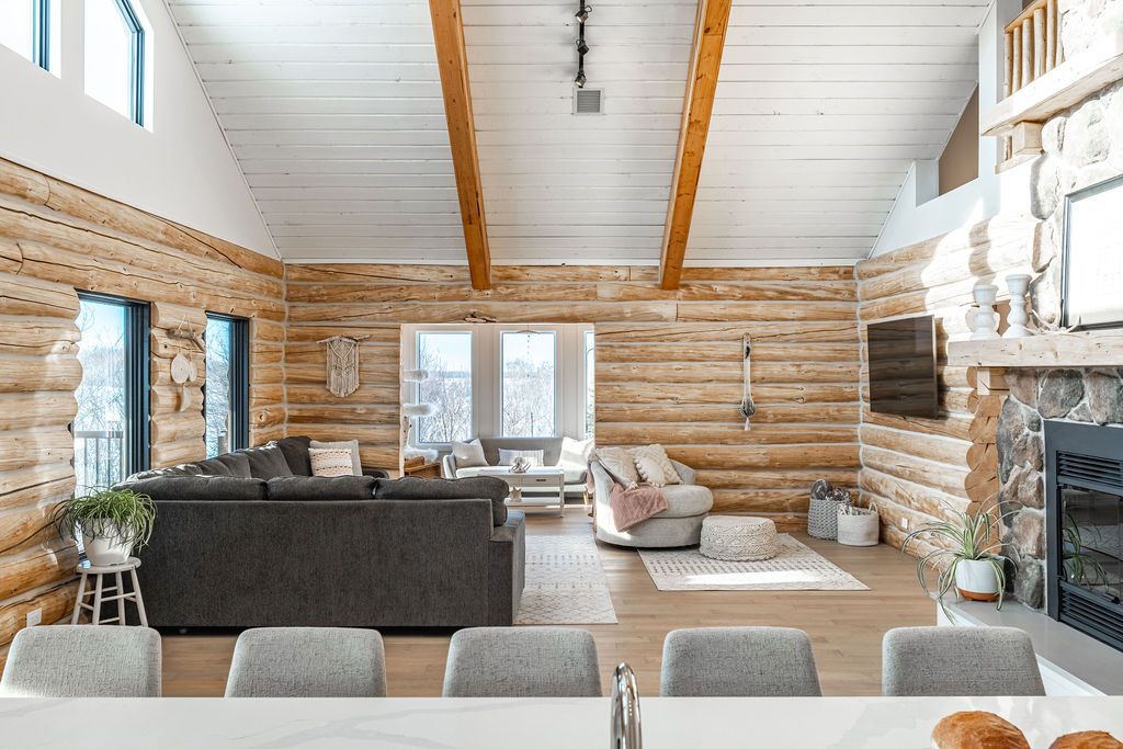 Bright, modern log cabin living room with exposed wood beams, large windows, gray and beige sofas, a stone fireplace, potted plants, and light wood flooring, viewed from a kitchen island.