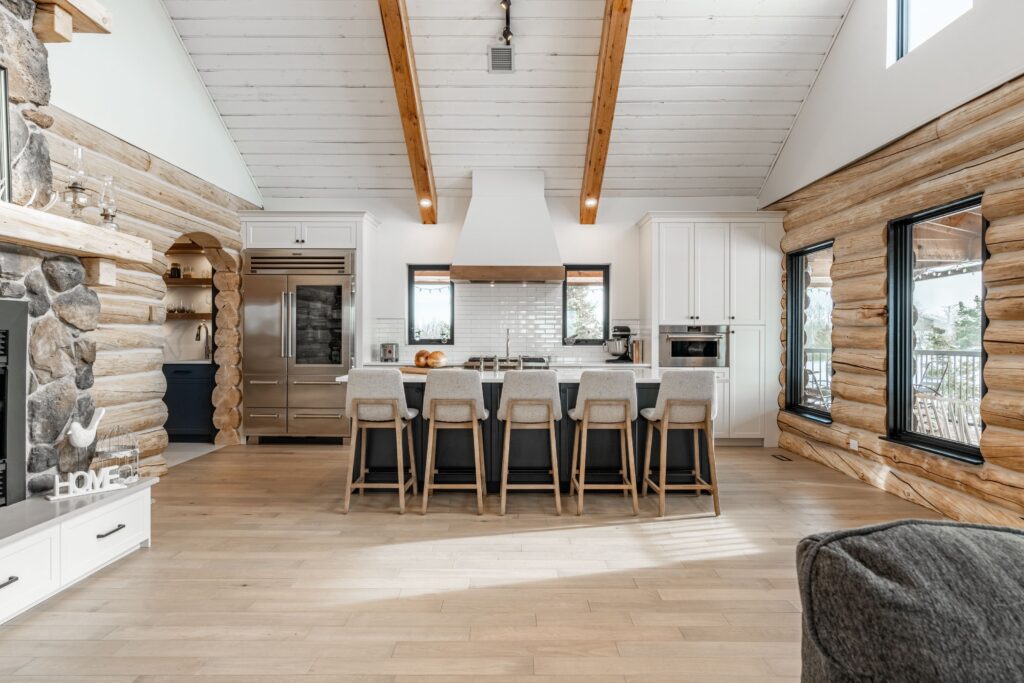 Spacious modern kitchen with a large island and five upholstered stools, white cabinetry, stainless steel appliances, wood beams, log walls, and light hardwood floors in a bright open-concept space.