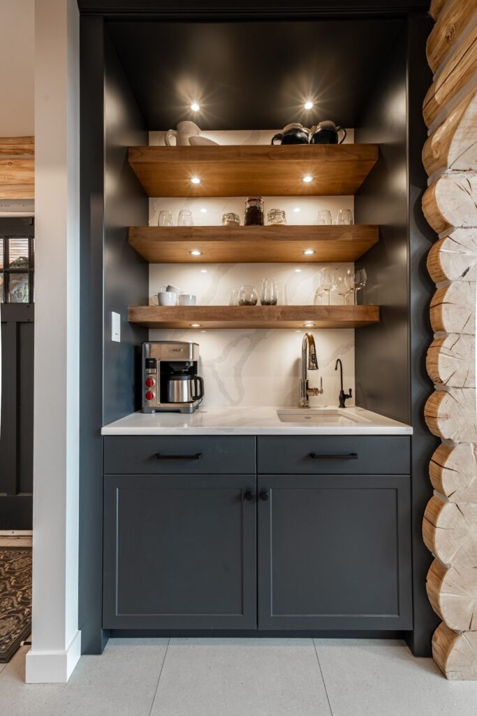 Modern coffee nook with dark cabinets, open wooden shelves holding cups and jars, a coffee maker on the white countertop, a small sink with a gold faucet, and accent lighting under each shelf.
