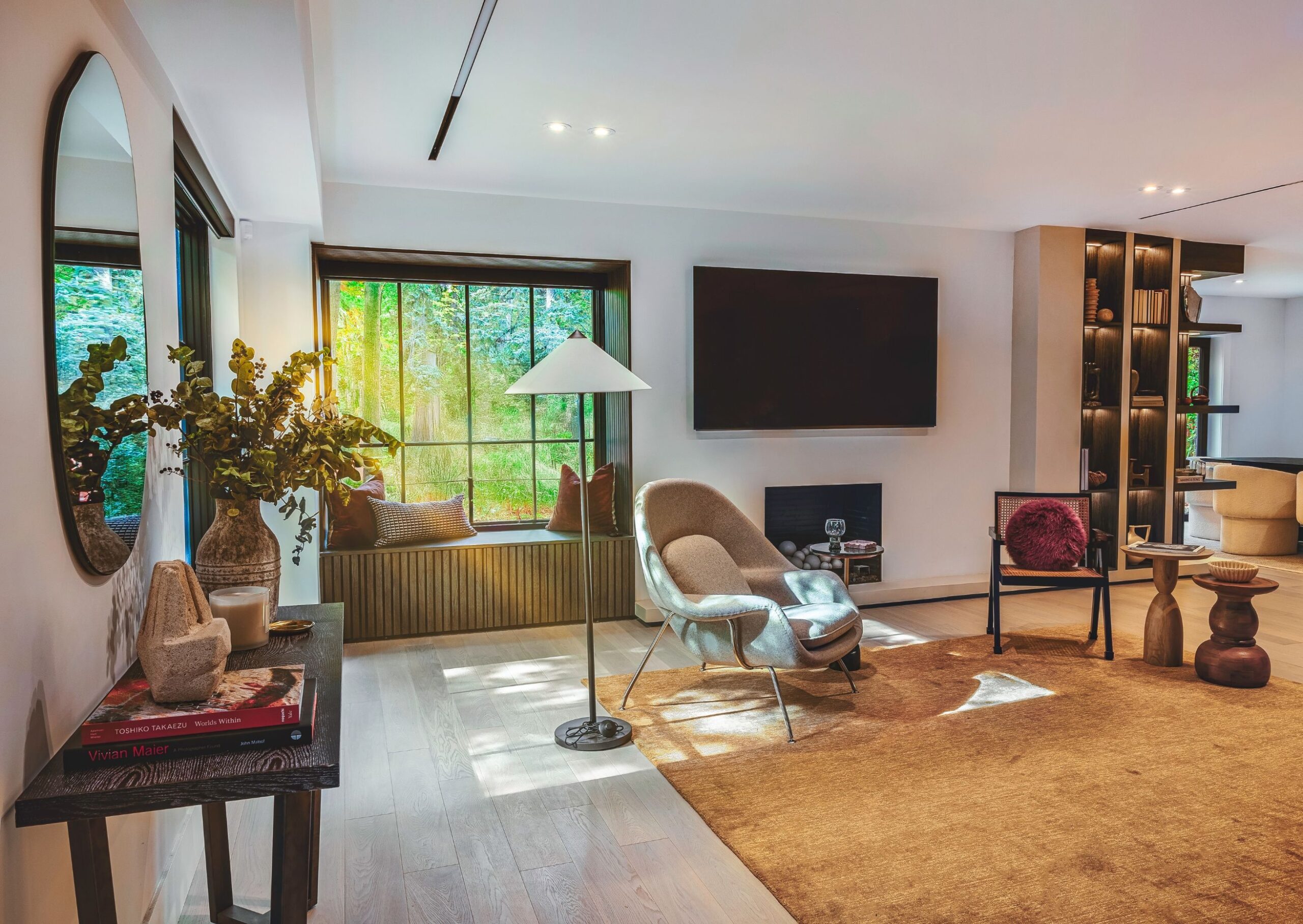 A modern living room with large windows, a cozy window seat, a flat-screen TV, a beige armchair with a floor lamp, shelves with decor, and a light brown rug on wooden flooring.