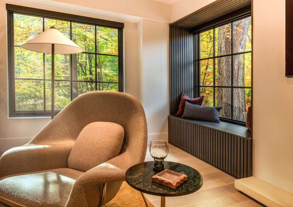 A cozy living room features a modern beige armchair, a round side table with a glass and book, large black-framed windows, a window seat with cushions, and vibrant autumn trees visible outside.
