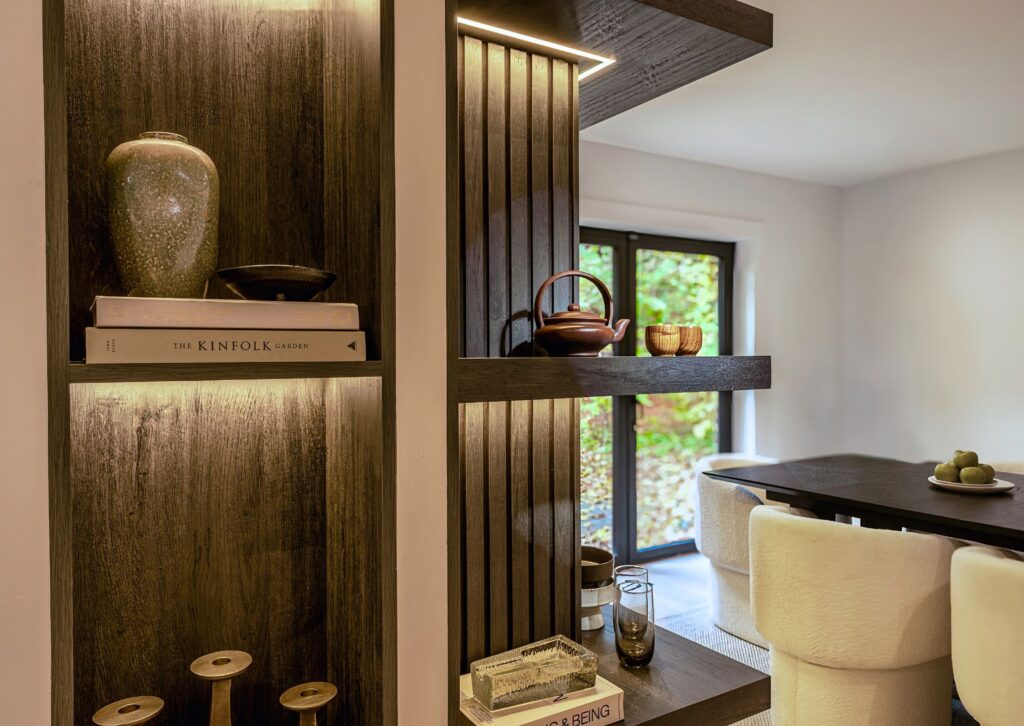 Modern wooden shelving unit with books, ceramics, and decorative items divides a cozy dining area with plush white chairs, a dark table, and large window overlooking greenery. Warm lighting highlights the shelf contents.