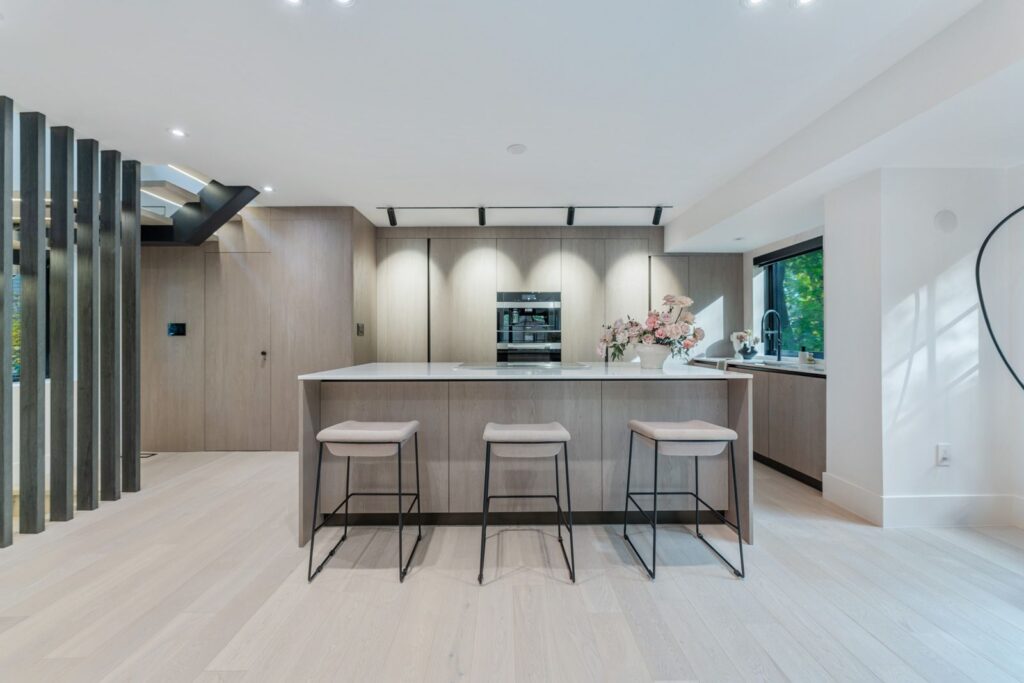 Modern kitchen with light wood cabinets, an island with three cushioned stools, built-in oven, minimalist decor, large windows, and bright natural lighting. Pink flowers and decorative items are arranged on the countertop.