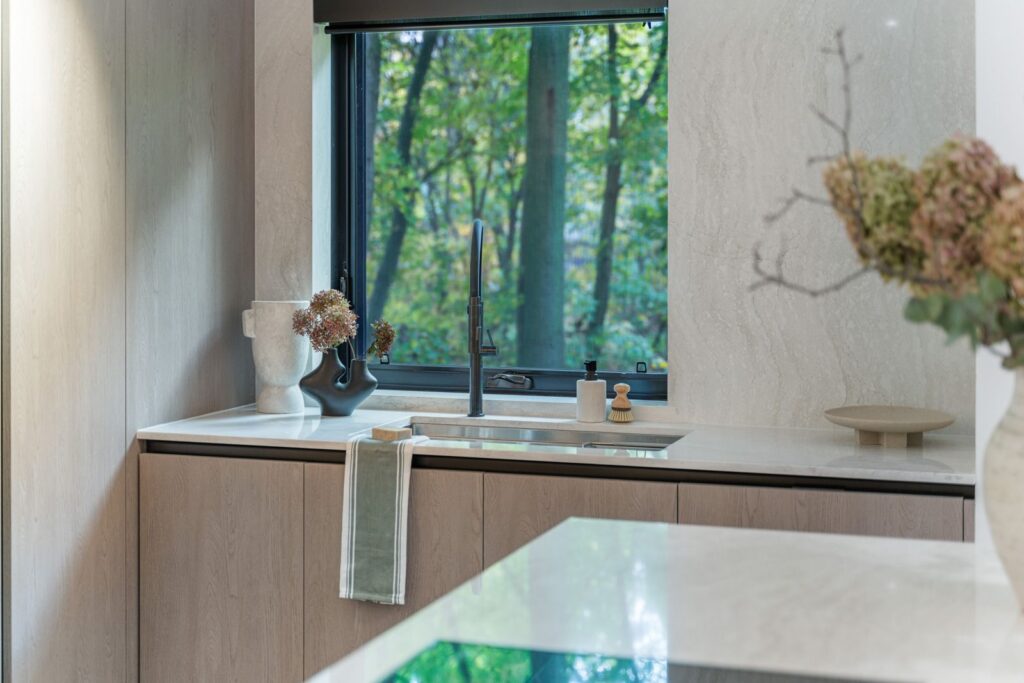 Modern kitchen with light wood cabinets and a marble countertop, featuring a sink under a window with a view of green trees. A towel hangs by the sink, and dried flowers decorate the counter.
