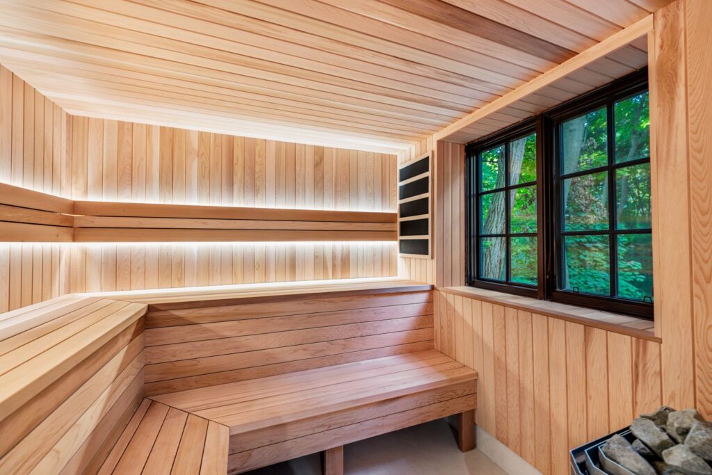 Modern wooden sauna with built-in benches, warm LED lighting, and a large window showing green foliage outside. A heater with sauna stones is visible in the bottom right corner.