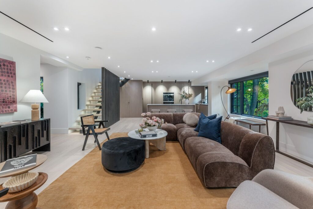 Modern open-concept living room with a curved brown sofa, round coffee tables, tan rug, and large windows. A staircase leads upstairs; the kitchen is visible in the background with sleek cabinets and built-in appliances.