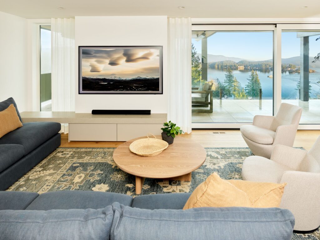 Modern living room with blue and beige furniture, a round wooden coffee table, TV displaying a scenic image, large window, and a view of trees, mountains, and water outside. Natural light fills the space.