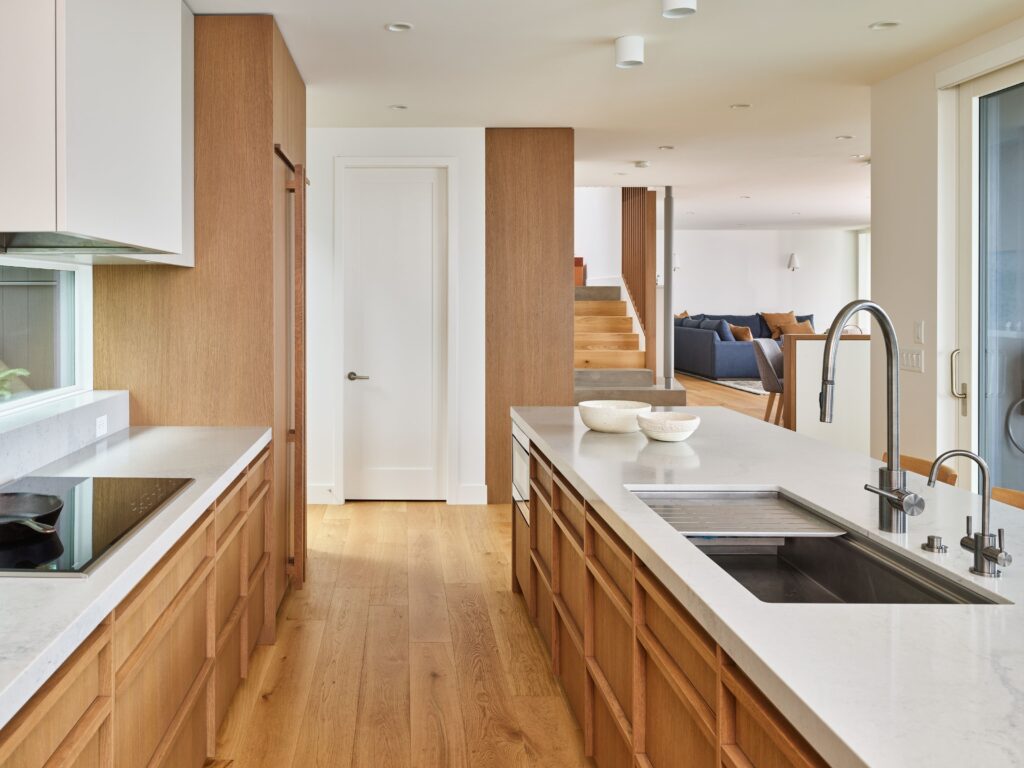 A modern kitchen with light wood cabinets, white countertops, a built-in stove, and a large island with a sink. The space is bright, with wooden floors and an open layout leading to a living area and stairs.