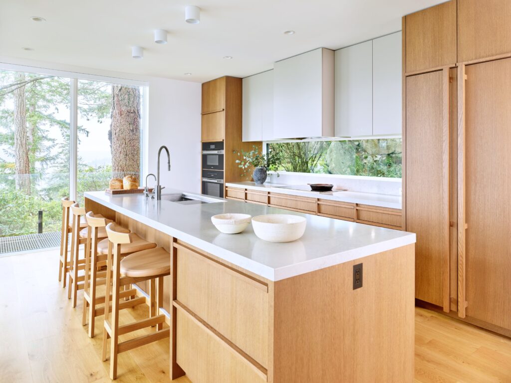 A modern kitchen with light wood cabinets, a large island with white countertop, four wooden barstools, built-in appliances, and large windows offering a view of trees outside.