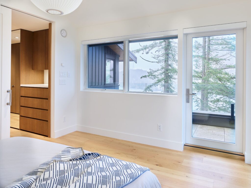 A bright, modern bedroom with light wood floors, white walls, and large windows overlooking trees and a balcony; a neatly made bed with a patterned blanket is partially visible.