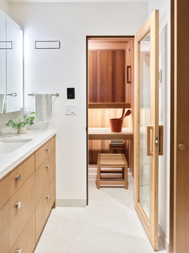 Modern bathroom with light wood cabinets and white counters; an open glass door leads to a wooden sauna with a small bench and a bucket, creating a clean and calming spa-like atmosphere.