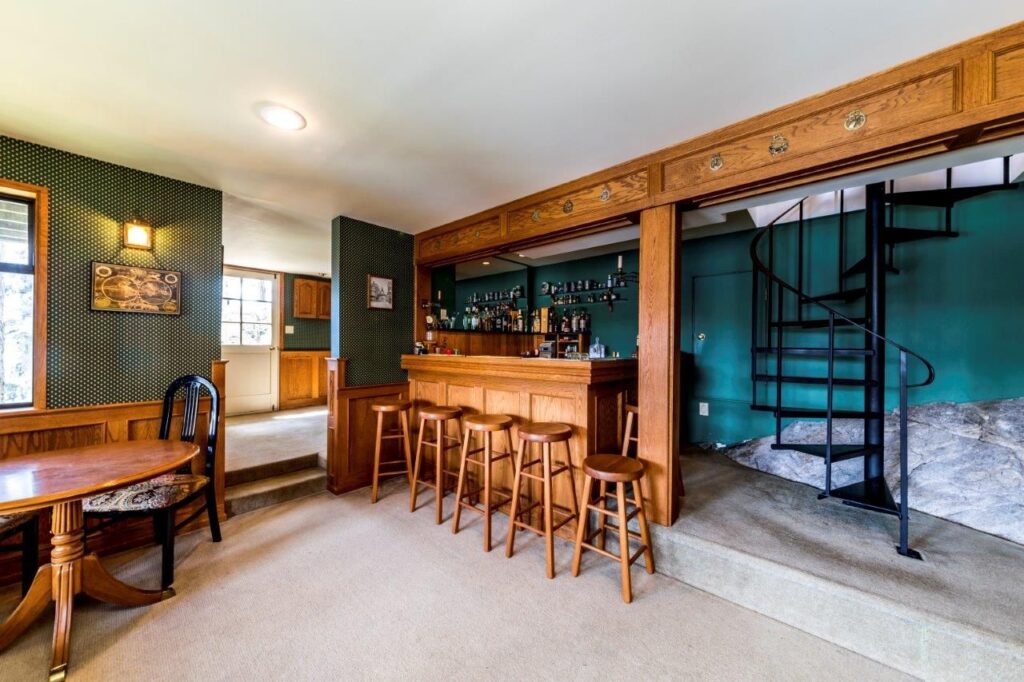 A home bar area with wooden stools, shelves of bottles, and a spiral staircase leading up. To the left, there is a round dining table with chairs and a window. The bar sits on a slightly elevated carpeted platform.