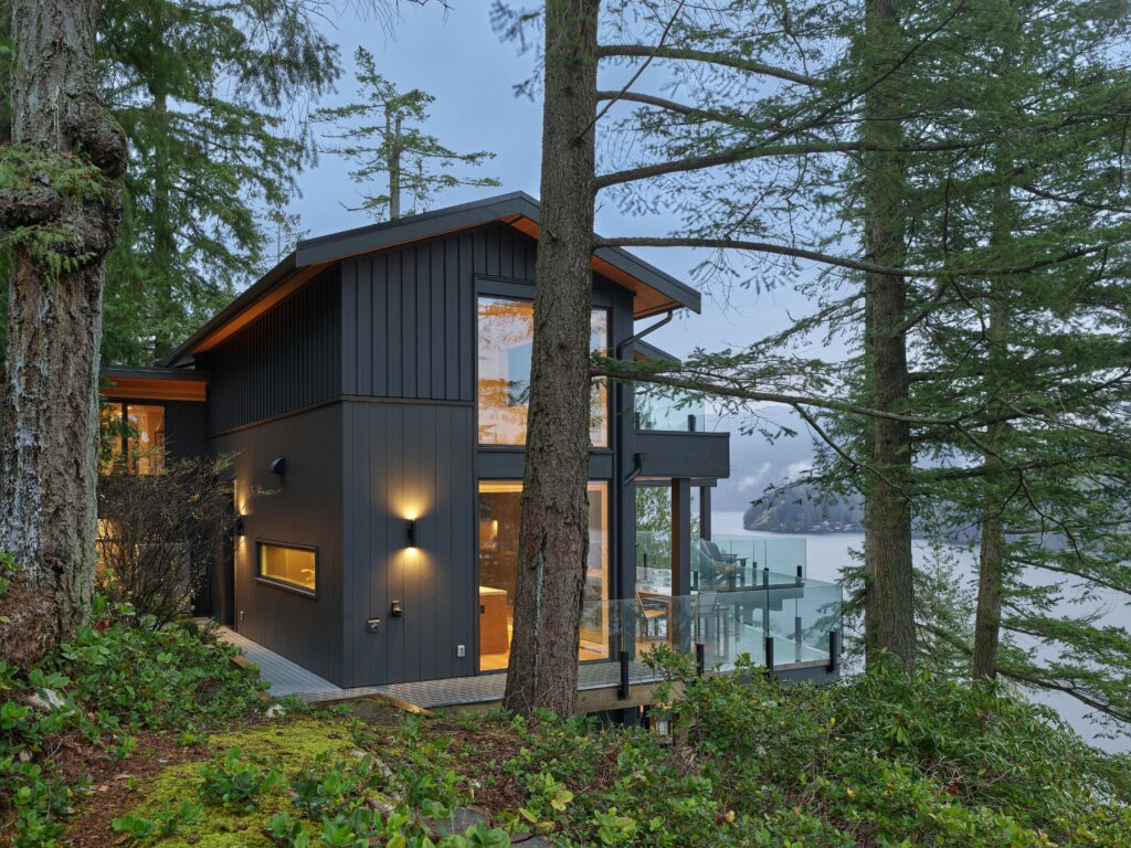 Modern two-story house with large windows and dark wood siding nestled among tall trees, overlooking a body of water and distant forested hills, creating a serene, natural setting.