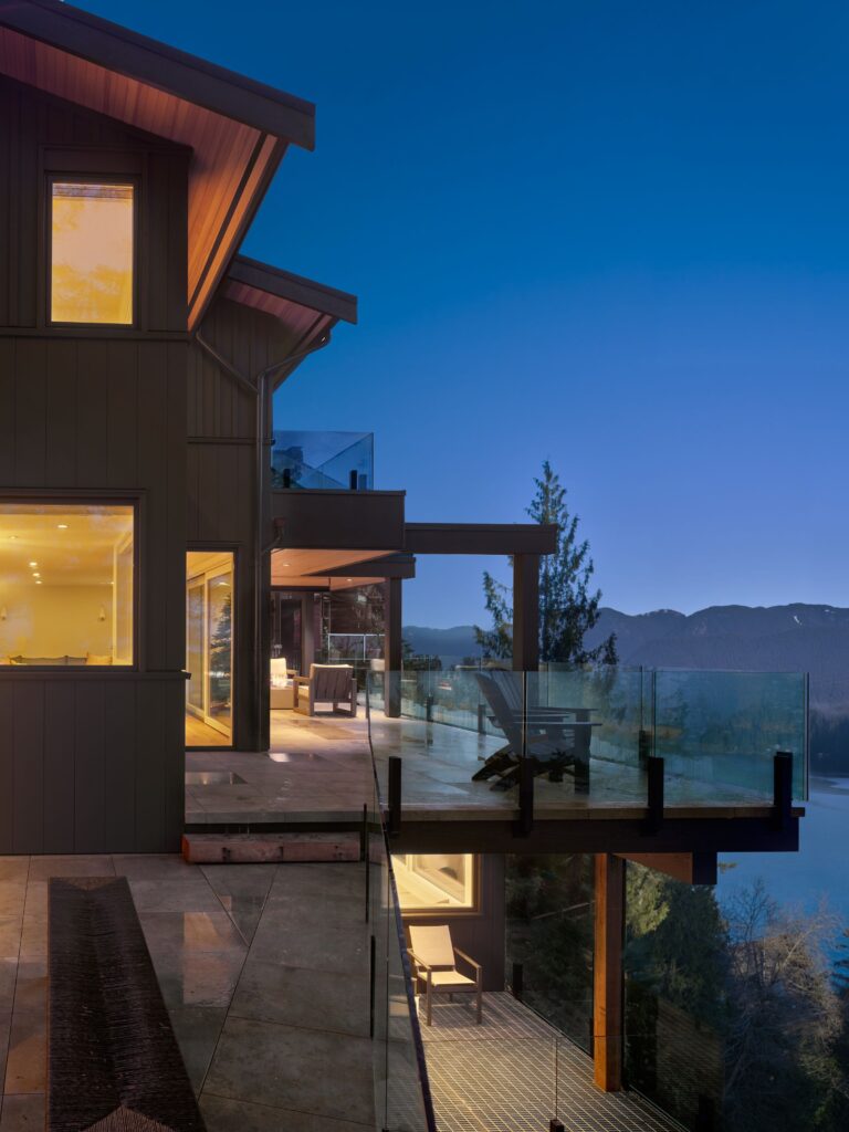 Modern house with large windows and glass railings overlooking a forested mountain landscape and lake at dusk, with warm interior lights contrasting the deep blue evening sky.