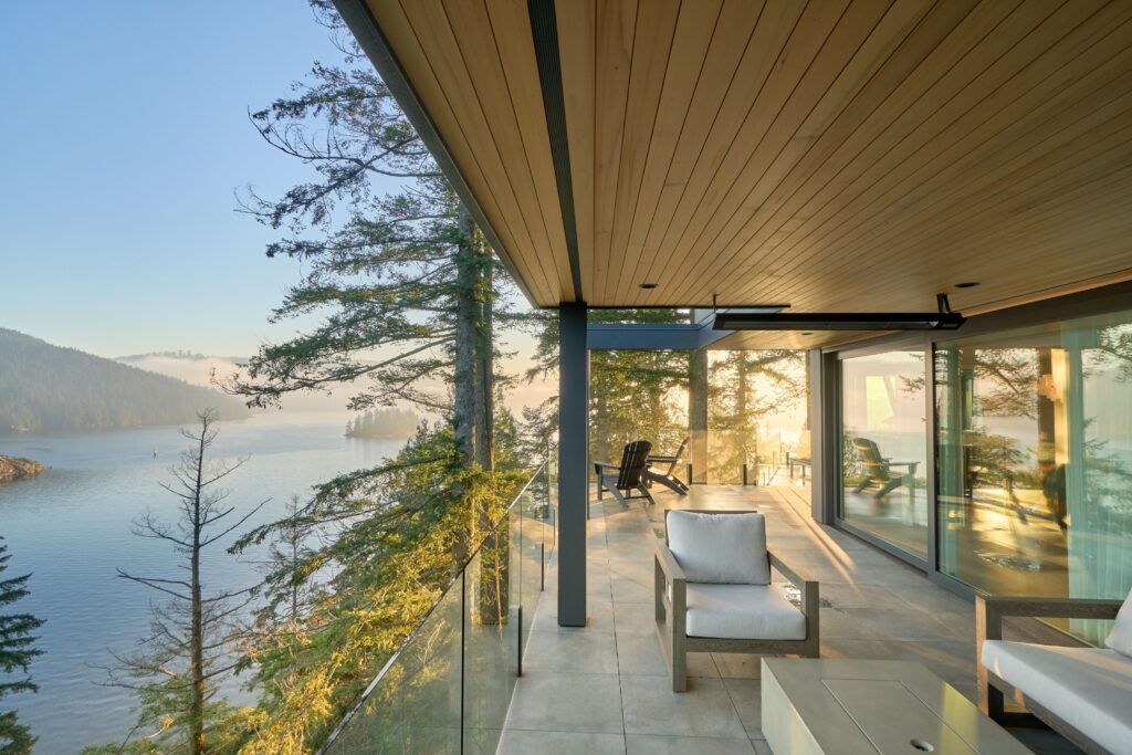 A modern balcony with glass railings and wooden ceiling overlooks a serene lake surrounded by trees and mountains. Outdoor seating faces the water, bathed in soft natural light.