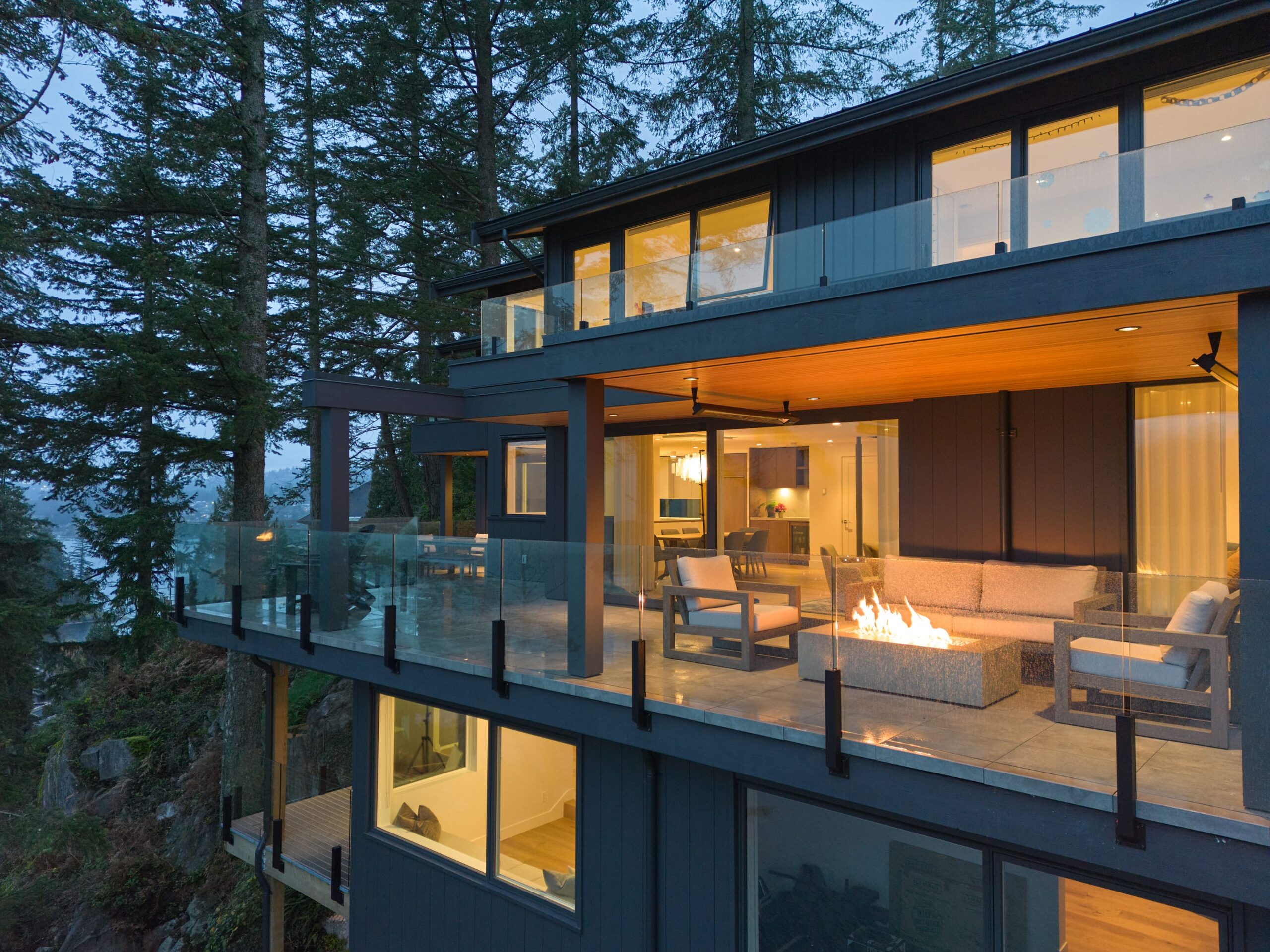 A modern multi-level house with large glass windows and balconies, surrounded by tall trees, features a cozy outdoor seating area with a fire pit glowing on the terrace at dusk.