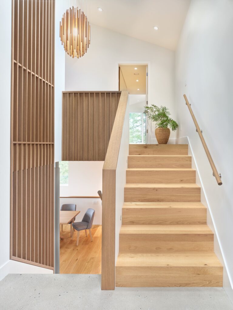 A bright, modern staircase with light wood steps and vertical wood slats. A potted plant sits at the top, and a wooden chandelier hangs above. Minimalist decor with white walls and natural light.