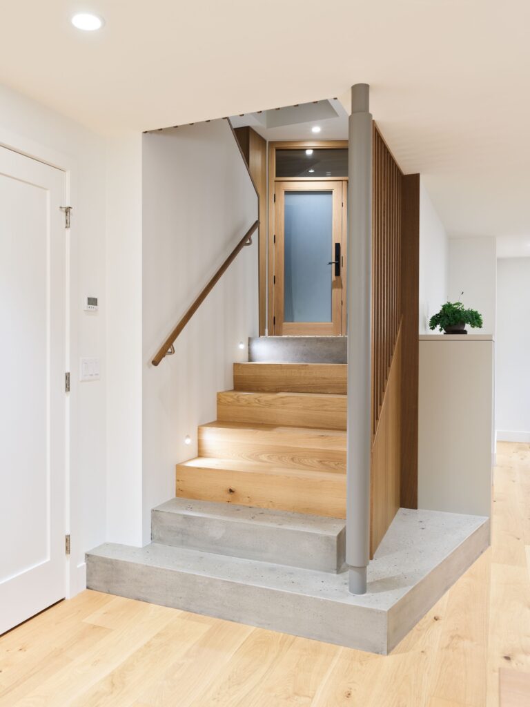 A modern staircase with wooden steps, concrete base, sleek railing, and built-in step lighting leads to a wooden door with frosted glass in a bright, minimalist interior.