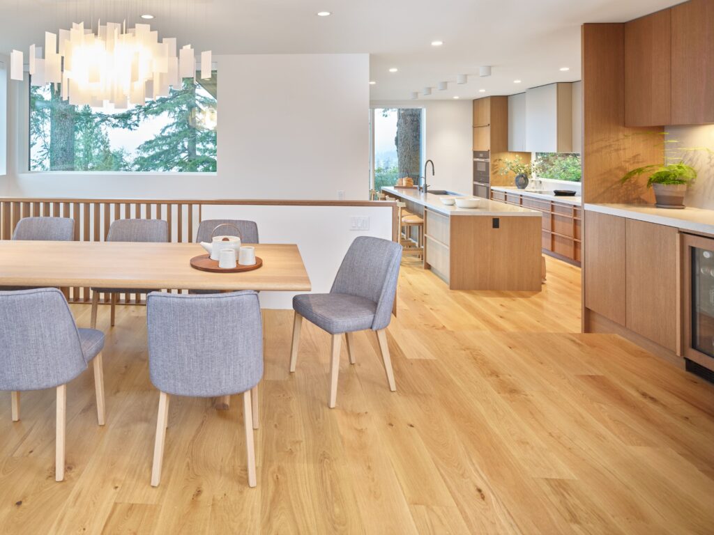 A modern dining and kitchen area with light wood floors, a wooden table with six gray chairs, a unique white chandelier, and a spacious kitchen featuring wood cabinets, marble countertops, and large windows with tree views.