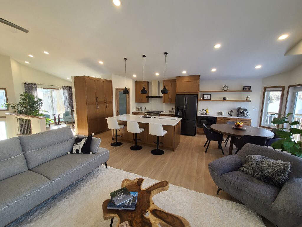 Modern open-concept kitchen and living area with wooden floors, gray sofa, armchair, white rug, unique wood coffee table, and kitchen island with pendant lights and barstools. Dining area and large windows let in natural light.