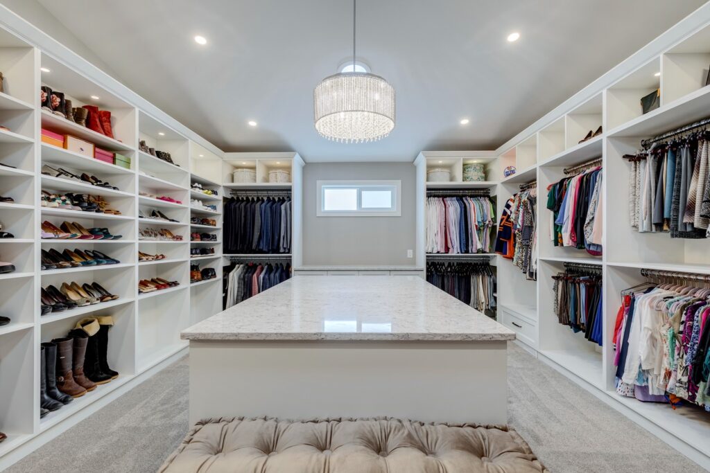 Spacious walk-in closet with built-in shelves and racks holding shoes, boots, clothing, and accessories. A marble-topped island sits in the center under a modern chandelier. A tufted bench is in the foreground.