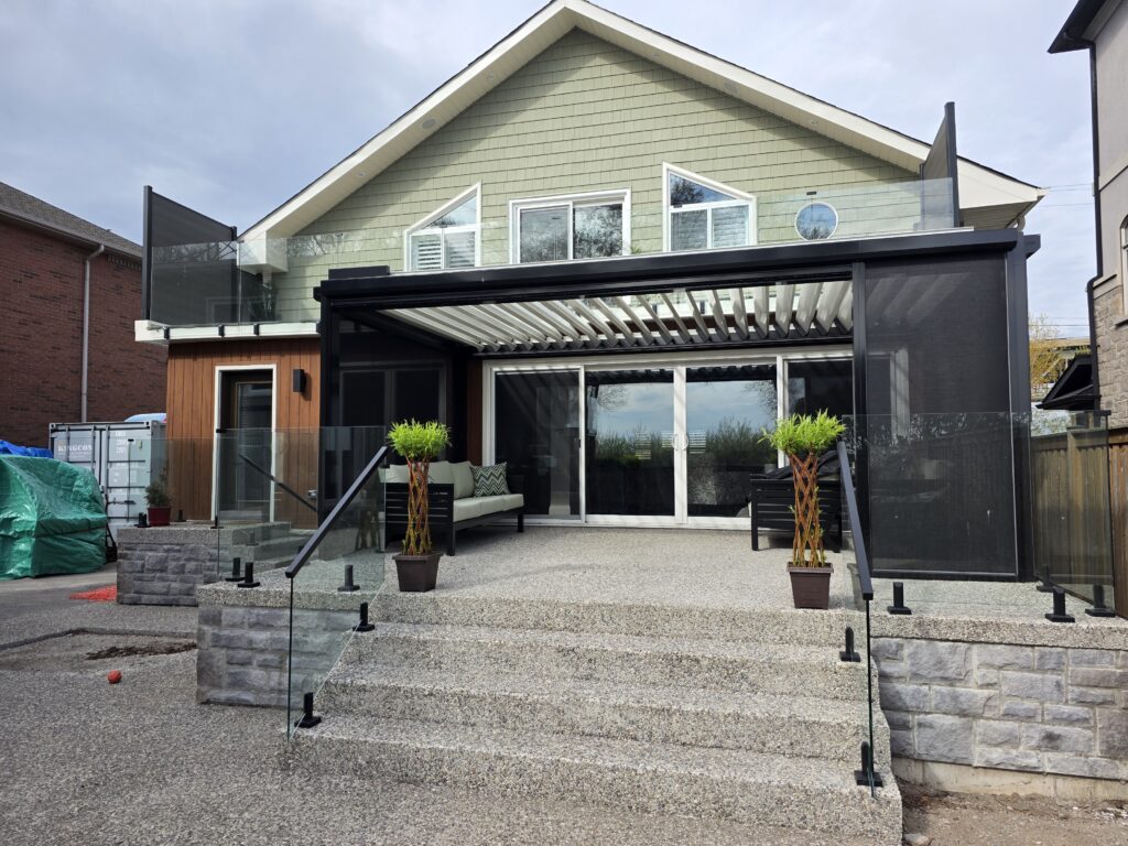 A modern two-story house with a large patio featuring a pergola, glass railings, potted plants, outdoor seating, and sliding glass doors. The house has beige siding and large windows on the upper floor.