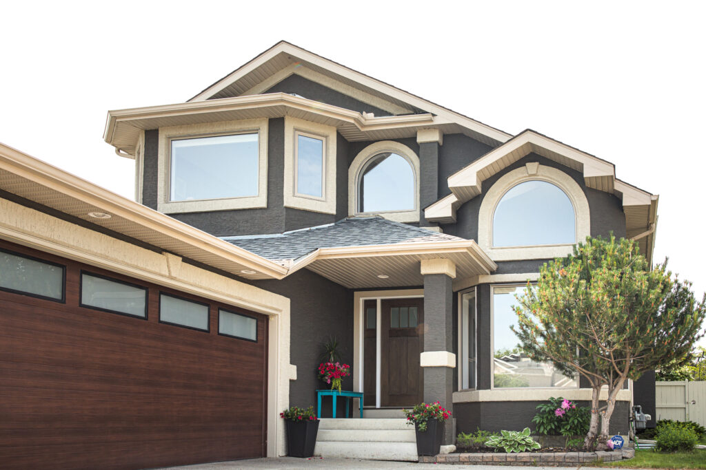 Two-story modern house with dark gray exterior, large windows, cream trim, and a wooden double garage door. A small front porch has potted plants, and a tree and flowers decorate the landscaped yard.