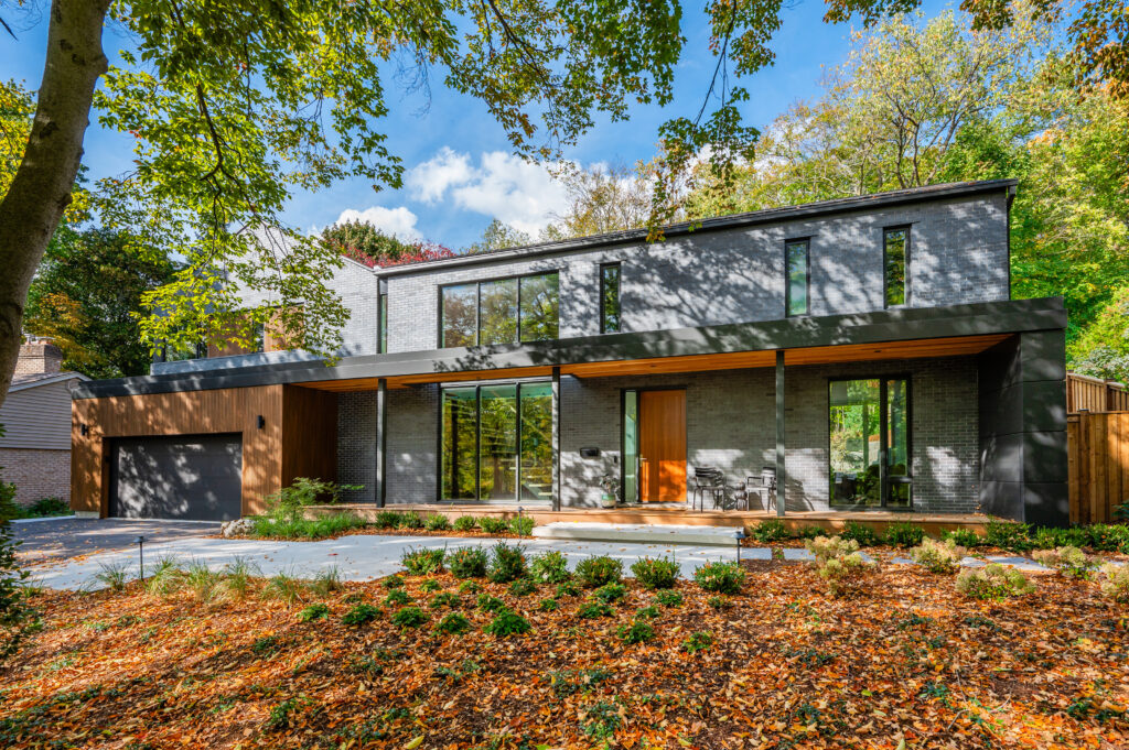Modern two-story house with large windows, gray brick exterior, and wooden accents, surrounded by trees and autumn leaves on the ground. A small patio area with chairs is visible near the entrance.