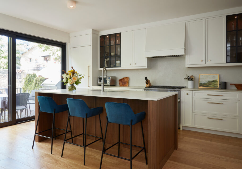 Modern kitchen with white cabinets, wood island, and three blue bar stools. A vase with flowers sits on the island. Large sliding glass doors let in natural light; outdoor patio and greenery are visible.