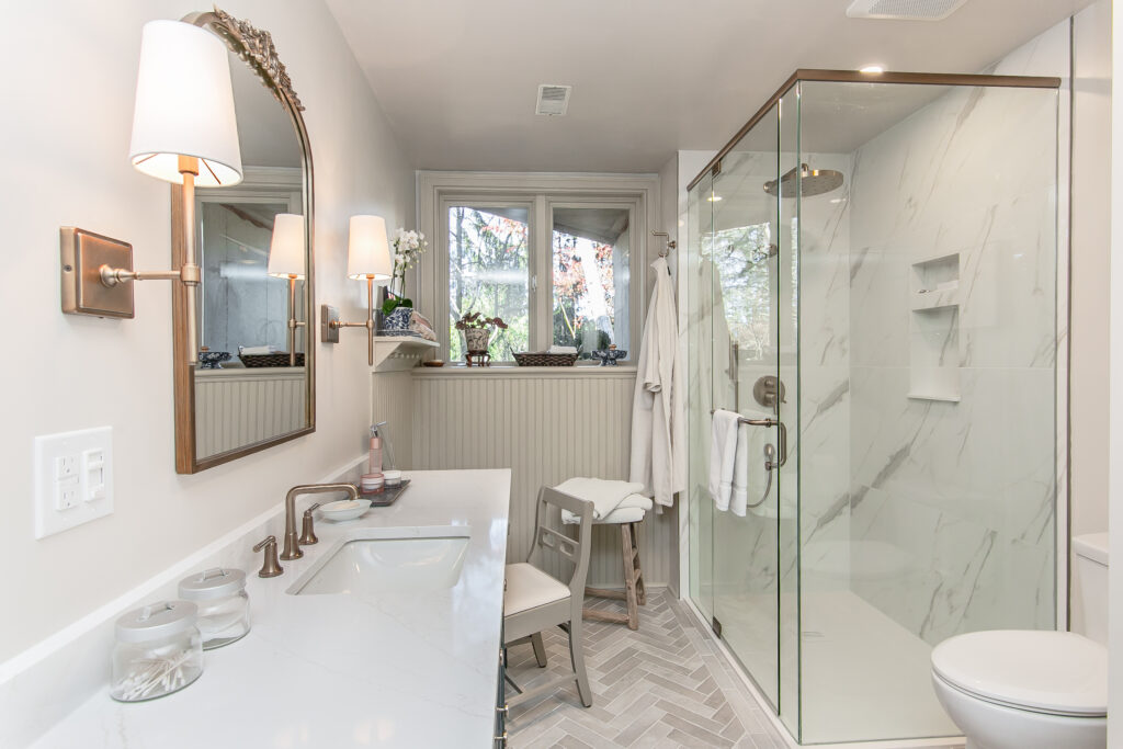 A modern bathroom with a double sink vanity, large mirror, glass shower, white walls, wall sconces, and a window above a small shelf with plants. A white stool and hanging robes are near the shower.