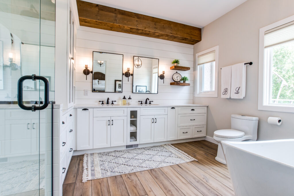 Bright, modern bathroom with double sinks, large mirror, white cabinets, glass shower, freestanding tub, wood floor, exposed wood beam, and decorative accents on shelves above the vanity. Natural light streams in from windows.