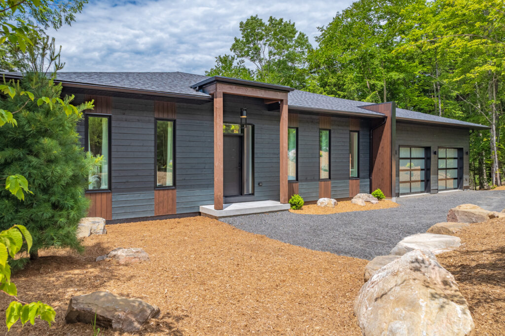 Modern single-story house with dark gray siding, large windows, wood accents, and a two-car garage with glass doors, surrounded by trees and landscaped with rocks and mulch.